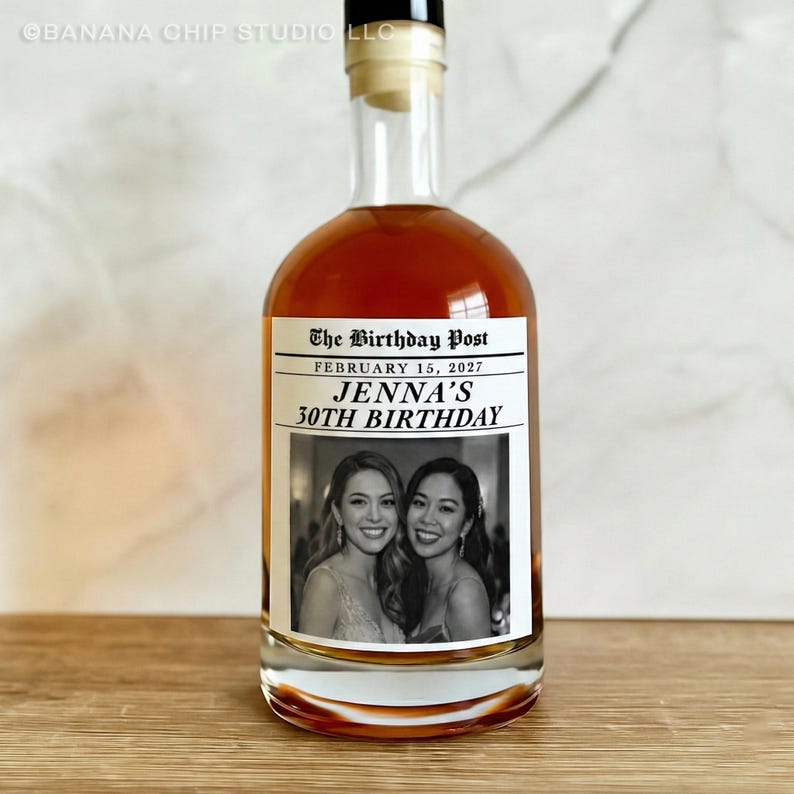 May include: A clear glass bottle filled with a brown liquid, likely a spirit. The label reads "The Birthday Post" with the date "February 15, 2027" and "Jenna's 30th Birthday." The label also features two women in a black and white photo.