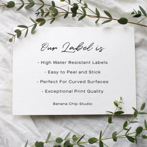 May include: A white card with the text "Our Label is" in elegant script. Below, bullet points list features: high water resistance, easy peel and stick, perfect for curved surfaces, and exceptional print quality. The card is surrounded by greenery.