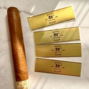 May include: Four gold-colored labels with black text that reads "Happy 21st Birthday Oliver 2004" and a brown cigar with a gold band.