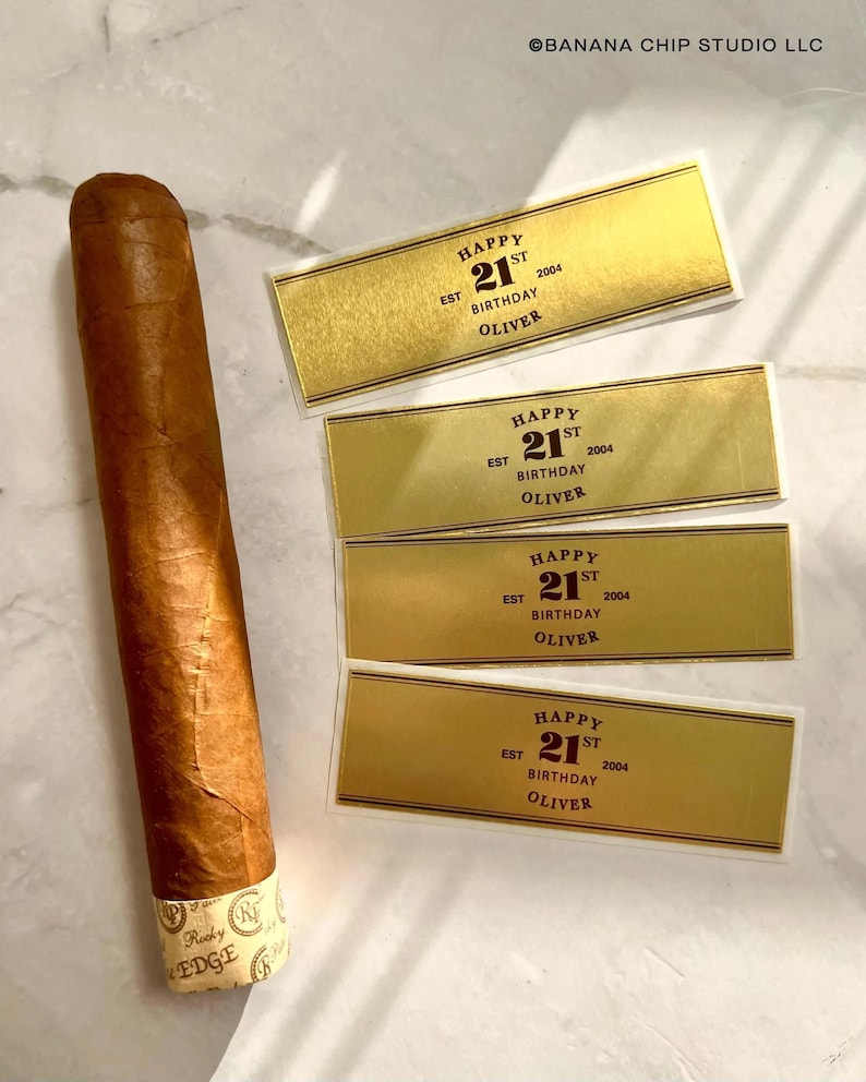 May include: Four gold-colored labels with black text that reads "Happy 21st Birthday Oliver 2004" and a brown cigar with a gold band.
