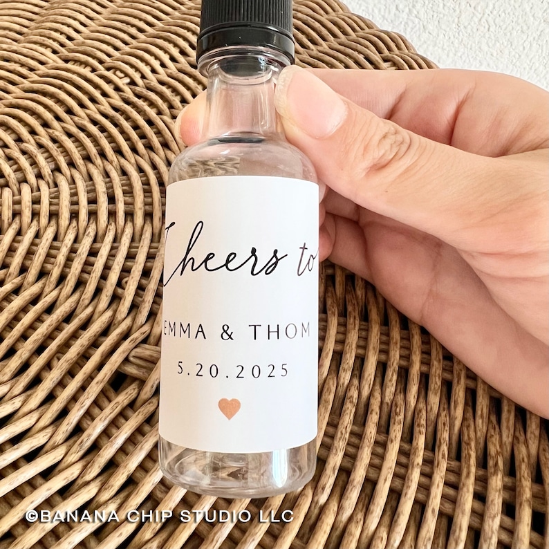 May include: A clear plastic bottle with a black screw cap featuring a white label. The label displays the black text Cheers to EMMA & THOM, the date 5.20.2025, and a small copper-colored heart icon centered at the bottom of the printed area.