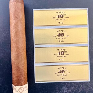 May include: Four gold-colored labels with black text that reads "Happy 40th Birthday Wil Est 1984". The labels are on a white background. A brown cigar is to the left of the labels.