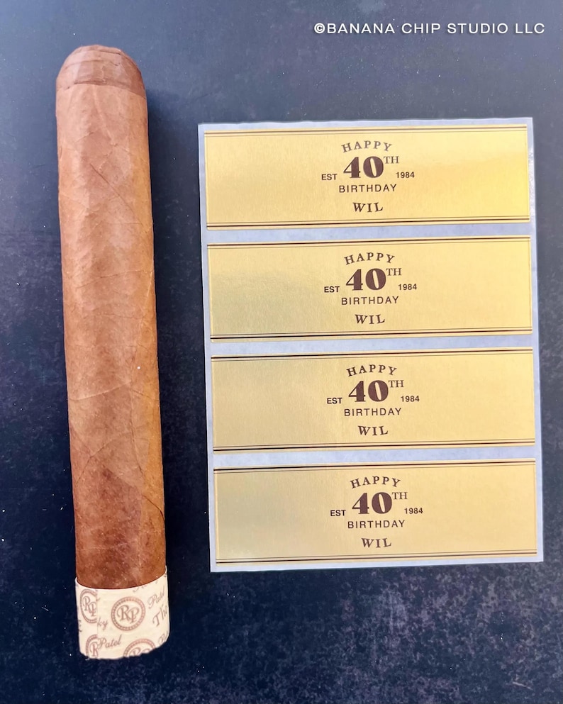 May include: Four gold-colored labels with black text that reads "Happy 40th Birthday Wil Est 1984". The labels are on a white background. A brown cigar is to the left of the labels.