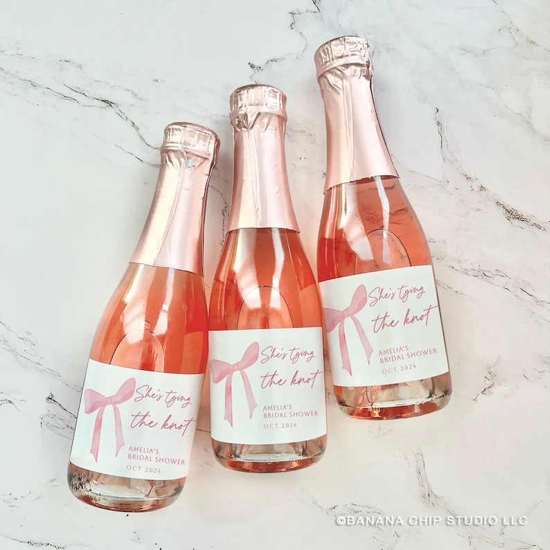 May include: Three miniature glass bottles filled with rose-colored liquid, each topped with a metallic pink foil cap. The white labels feature a pink bow illustration and the text: She's tying the knot, AMELIA'S BRIDAL SHOWER, OCT 2024.