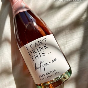 以下が含まれることがあります： 白いラベルが付いた、バラ色のシャンパンボトル。「I CAN'T DRINK THIS... but you can」と「BABY AMELIA DUE APRIL 2026」と書かれています。 ボトルは10.16cm x 8.26cmで、750mlのシャンパンが入っています。