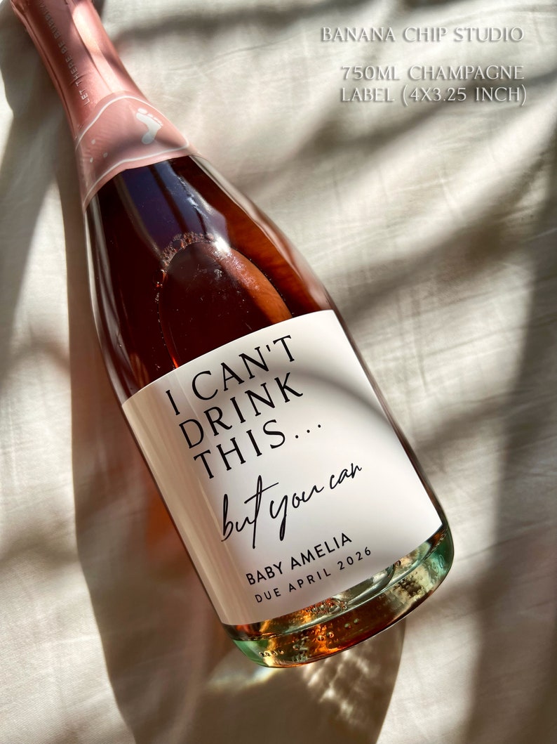 以下が含まれることがあります： 白いラベルが付いた、バラ色のシャンパンボトル。「I CAN'T DRINK THIS... but you can」と「BABY AMELIA DUE APRIL 2026」と書かれています。 ボトルは10.16cm x 8.26cmで、750mlのシャンパンが入っています。