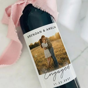 May include: A dark glass wine bottle with a custom label featuring a couple embracing in a field. The label reads "Brendan & Amelia" and "Engaged 11.22.2027." A pink ribbon is tied around the bottle's neck.