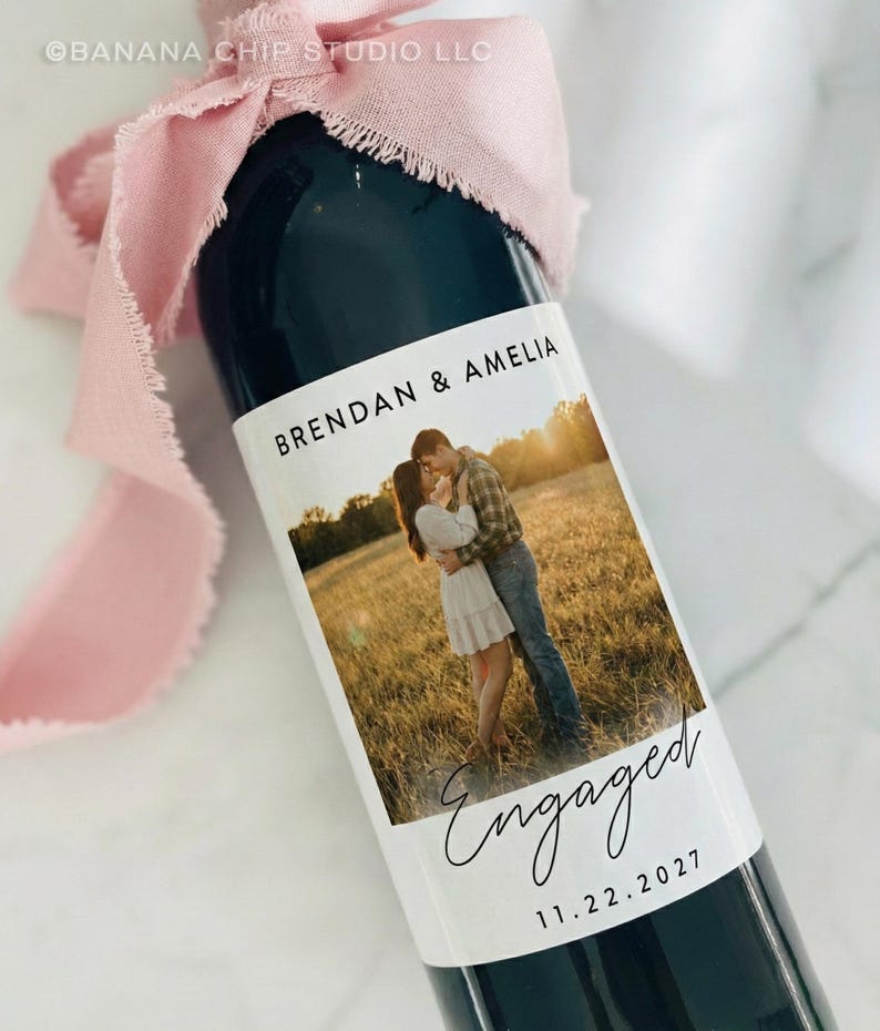 May include: A dark glass wine bottle with a custom label featuring a couple embracing in a field. The label reads "Brendan & Amelia" and "Engaged 11.22.2027." A pink ribbon is tied around the bottle's neck.