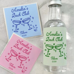 May include: A clear glass miniature bottle with a silver cap and a light green label featuring the text Amelia's Bach Club, Tulum, 2025, and two glasses. Two square stickers in light blue and pink with identical text and graphics are included.