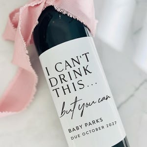 以下が含まれることがあります： 「I CAN'T DRINK THIS... but you can」と黒字で書かれた白いラベルのワインボトル。ボトルはピンクのリボンで飾られています。ラベルには「BABY PARKS DUE OCTOBER 2027」とも書かれています。