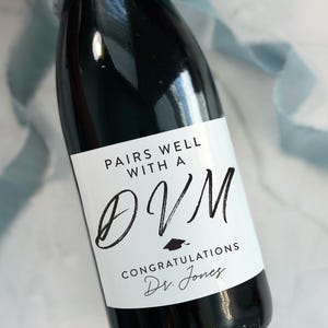 May include: A black champagne bottle with a white label that reads "PAIRS WELL WITH A DVM CONGRATULATIONS Dr. Jones." The bottle is tied with a light blue ribbon. The bottle is 4 inches by 3.25 inches.