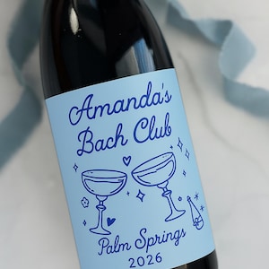 May include: A dark glass bottle with a light blue label measuring 3.5x4 inches. The label features dark blue text reading Amanda's Bach Club, Palm Springs 2026, with illustrations of two champagne coupes, a bottle, hearts, and stars.