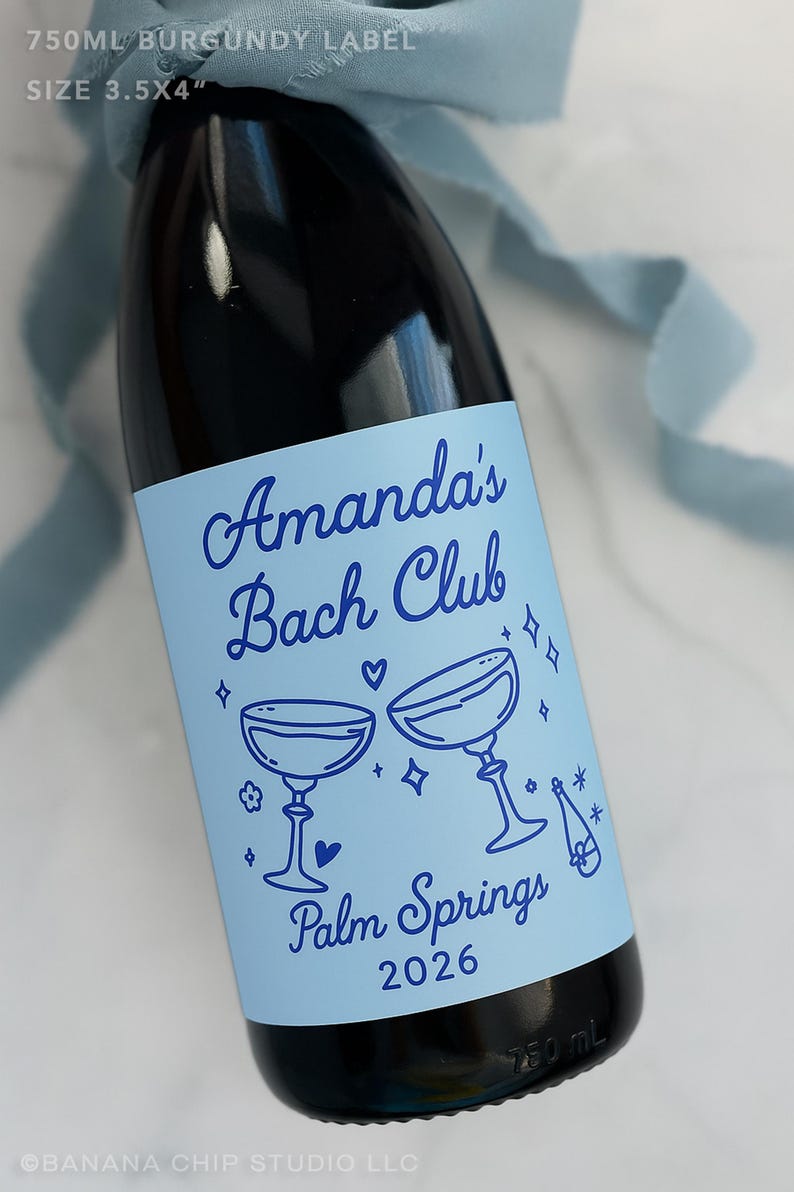 May include: A dark glass bottle with a light blue label measuring 3.5x4 inches. The label features dark blue text reading Amanda's Bach Club, Palm Springs 2026, with illustrations of two champagne coupes, a bottle, hearts, and stars.
