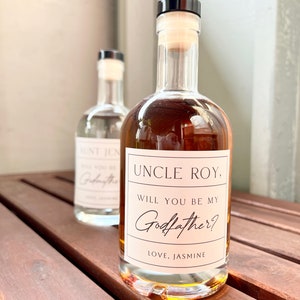 May include: Two glass bottles with cork stoppers. The front bottle has a label reading "UNCLE ROY, WILL YOU BE MY Godfather? LOVE, JASMINE." The second bottle has a label reading "AUNT JEN, WILL YOU BE MY Godfather?"