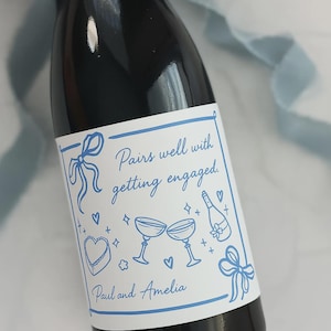 May include: A dark wine bottle with a white label featuring blue illustrations of champagne glasses, hearts, and a bow. The label reads "Pairs well with getting engaged" and includes the names "Paul and Amelia".