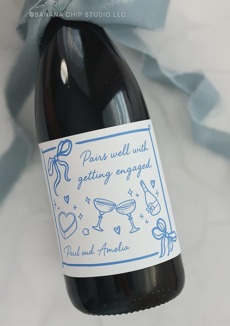May include: A dark wine bottle with a white label featuring blue illustrations of champagne glasses, hearts, and a bow. The label reads "Pairs well with getting engaged" and includes the names "Paul and Amelia".