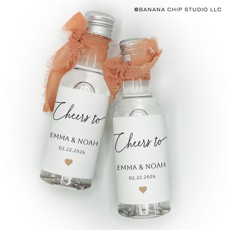 May include: Two clear glass mini bottles with silver screw caps, each tied with a frayed peach-colored ribbon. The white labels feature the black text Cheers to EMMA & NOAH 02.22.2026 and a small gold heart icon. The text BANANA CHIP STUDIO LLC is visible.