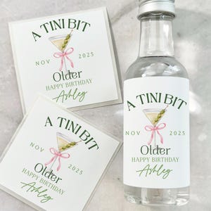 Puede incluir: Una botella de vidrio transparente con tapa plateada y una etiqueta que dice "A Tini Bit Older Happy Birthday Ashley" con una ilustraci&oacute;n de martini. Dos tarjetas a juego est&aacute;n junto a la botella.