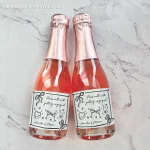 May include: Two mini champagne bottles with pink foil tops and labels that read "Pairs well with getting engaged." The labels feature bow and heart illustrations, and the names "Amelia & Mason." The bottles are filled with a pink liquid.