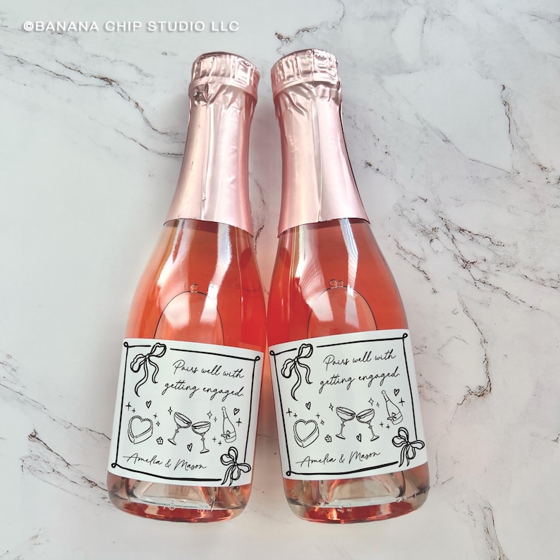 May include: Two mini champagne bottles with pink foil tops and labels that read "Pairs well with getting engaged." The labels feature bow and heart illustrations, and the names "Amelia & Mason." The bottles are filled with a pink liquid.