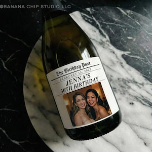 May include: A champagne bottle with a custom label. The label reads "The Birthday Post" with the date February 15, 2027, and "Jenna's 30th Birthday." The label also features two smiling women. The bottle is on a marble surface.