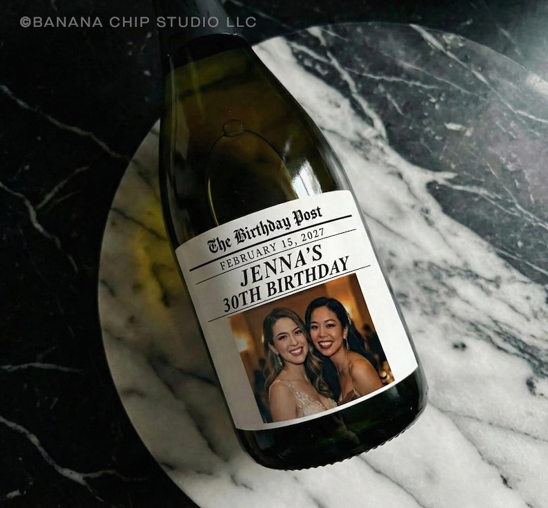 May include: A champagne bottle with a custom label. The label reads "The Birthday Post" with the date February 15, 2027, and "Jenna's 30th Birthday." The label also features two smiling women. The bottle is on a marble surface.