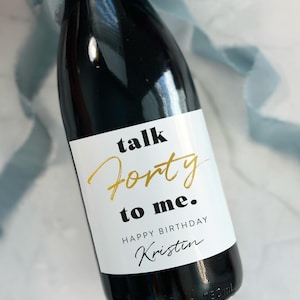 May include: A dark glass bottle with a white label featuring the black and gold text "talk Forty to me. HAPPY BIRTHDAY Kristin". The label measures 4 by 3.25 inches. A dusty blue ribbon is tied around the neck of the bottle.