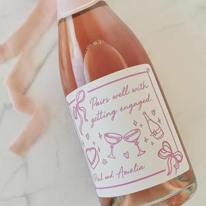 May include: A bottle of ros&eacute; wine with a custom label that reads "Pairs well with getting engaged." The label features pink illustrations of champagne glasses, hearts, and bows. A pink ribbon is draped near the bottle.