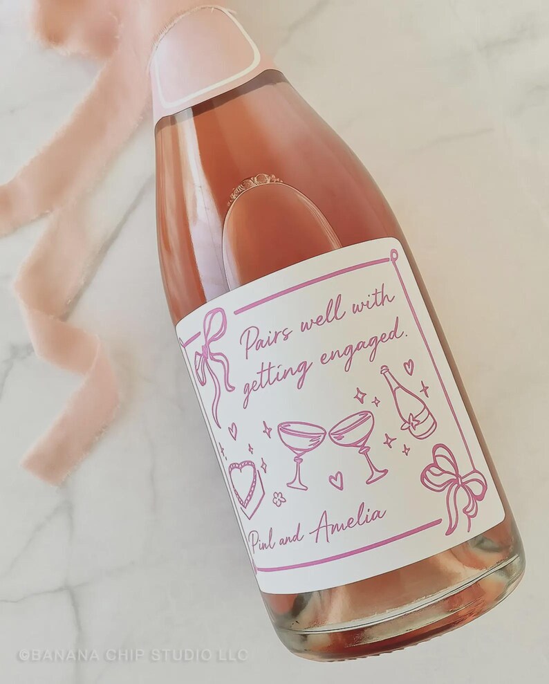 May include: A bottle of ros&eacute; wine with a custom label that reads "Pairs well with getting engaged." The label features pink illustrations of champagne glasses, hearts, and bows. A pink ribbon is draped near the bottle.