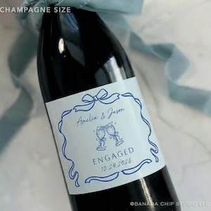 Puede incluir: Una botella de champ&aacute;n oscura con una etiqueta blanca con texto e ilustraciones azules. La etiqueta dice "Amelia & Jason" encima de dos copas de champ&aacute;n brindando, con "ENGAGED 10.24.2026" debajo. Una cinta azul est&aacute; atada alrededor de la botella.