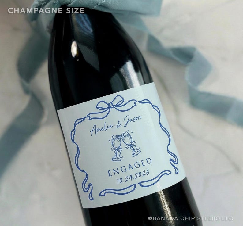 Puede incluir: Una botella de champ&aacute;n oscura con una etiqueta blanca con texto e ilustraciones azules. La etiqueta dice "Amelia & Jason" encima de dos copas de champ&aacute;n brindando, con "ENGAGED 10.24.2026" debajo. Una cinta azul est&aacute; atada alrededor de la botella.