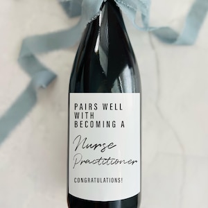 May include: A dark wine bottle with a white label that reads "Pairs Well With Becoming A Nurse Practitioner Congratulations!". The bottle is tied with a light blue ribbon. The label is 8.9x10.2 cm.