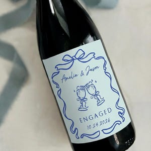 Puede incluir: Una botella de vino burdeos oscuro con una etiqueta personalizada. La etiqueta es azul claro con un dise&ntilde;o de cinta azul, los nombres "Amelia & Jason" y la palabra "ENGAGED" con la fecha 10.24.2026.
