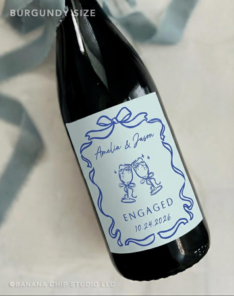Puede incluir: Una botella de vino burdeos oscuro con una etiqueta personalizada. La etiqueta es azul claro con un dise&ntilde;o de cinta azul, los nombres "Amelia & Jason" y la palabra "ENGAGED" con la fecha 10.24.2026.