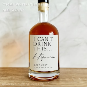 以下が含まれることがあります： 白いラベルが付いた透明なガラスのウイスキーボトル。ラベルには「I CAN'T DRINK THIS... but you can」と書かれており、その下に「BABY GIBBY DUE MARCH 202X」と書かれています。ボトルには琥珀色の液体が入っています。
