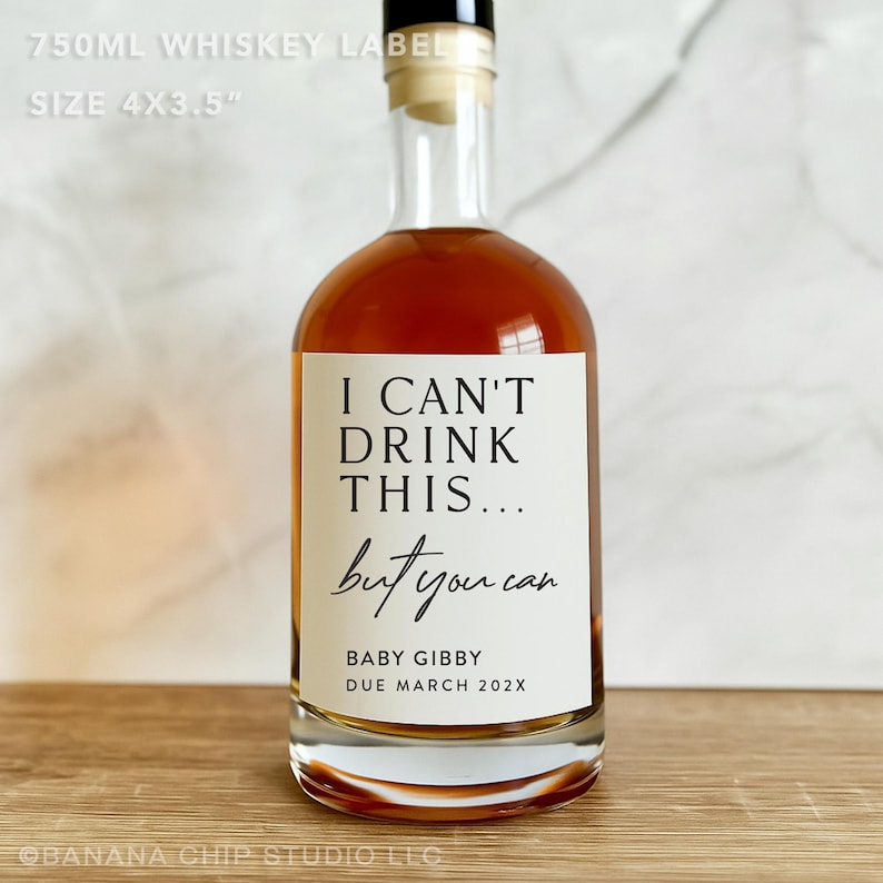 以下が含まれることがあります： 白いラベルが付いた透明なガラスのウイスキーボトル。ラベルには「I CAN'T DRINK THIS... but you can」と書かれており、その下に「BABY GIBBY DUE MARCH 202X」と書かれています。ボトルには琥珀色の液体が入っています。