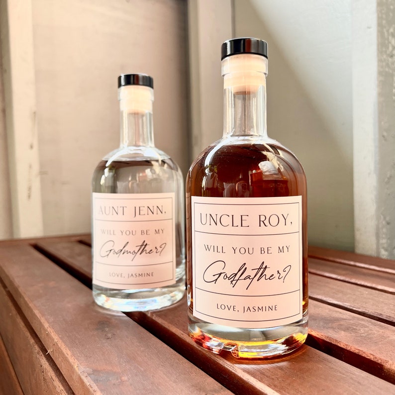 May include: Two glass bottles with black caps and cork stoppers. One bottle contains a clear liquid, the other a brown liquid. Each bottle has a pink label with the text "AUNT JENN, WILL YOU BE MY Godmother? LOVE, JASMINE" and "UNCLE ROY, WILL YOU BE MY Godfather? LOVE, JASMINE".