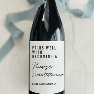 May include: A dark wine bottle with a white label that reads "Pairs Well With Becoming a Nurse Practitioner." The bottle is tied with a light blue ribbon. The label is 8.89 x 10.16 cm.