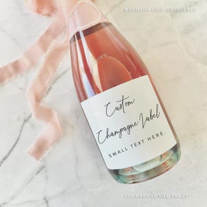 May include: A champagne bottle with a custom label. The label reads "Custom Champagne Label" with the text "SMALL TEXT HERE." A pink ribbon is tied around the neck of the bottle. The bottle is 4 x 3.25 inches.