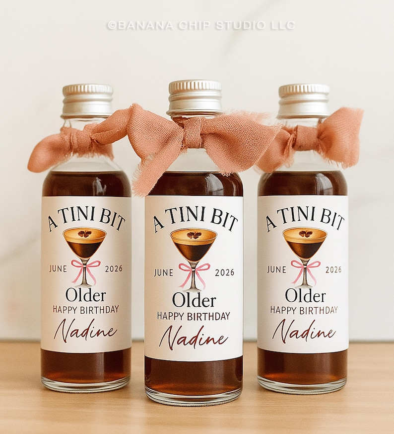 Puede incluir: Tres peque&ntilde;as botellas de vidrio llenas de un l&iacute;quido oscuro, cada una con una tapa plateada y una cinta color melocot&oacute;n atada alrededor del cuello. Las etiquetas dicen "A TINI BIT" con una ilustraci&oacute;n de copa de martini, "Junio 2026", "Older Happy Birthday Nadine".