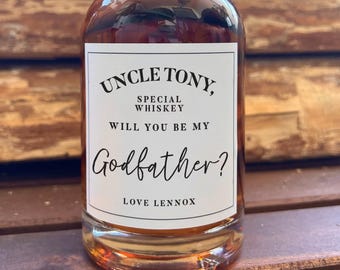 May include: A clear glass bottle filled with amber liquid featuring a white rectangular label with black text that reads: UNCLE TONY, SPECIAL WHISKEY WILL YOU BE MY Godfather? LOVE LENNOX. The bottle has a rounded shoulder and a short neck.