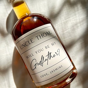 May include: A glass bottle filled with amber liquid, with a label saying "UNCLE THOM, WILL YOU BE MY Godfather? LOVE, JASMINE." The bottle has a black stopper and is approximately 7.6 cm by 7.6 cm.