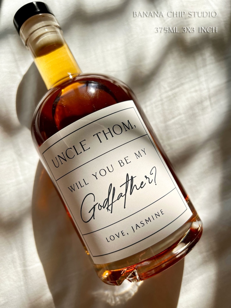 May include: A glass bottle filled with amber liquid, with a label saying "UNCLE THOM, WILL YOU BE MY Godfather? LOVE, JASMINE." The bottle has a black stopper and is approximately 7.6 cm by 7.6 cm.