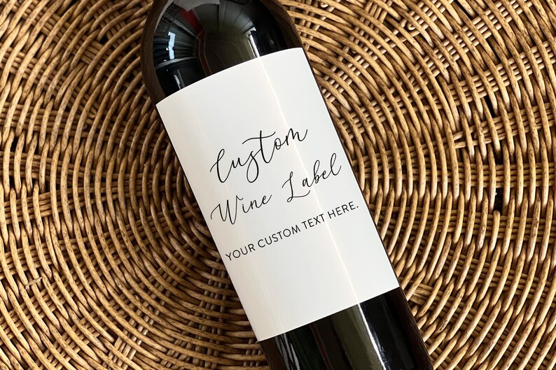 May include: A wine bottle with a white label that reads "Custom Wine Label" in elegant script. The bottom of the label says "YOUR CUSTOM TEXT HERE." The bottle is set against a woven, light brown background.