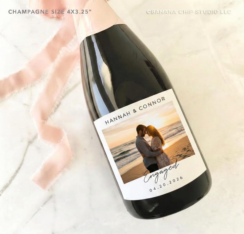 May include: A champagne bottle with a custom label featuring a couple embracing on a beach at sunset. The label reads "Hannah & Connor" and "Engaged 04.20.2026". The bottle is adorned with a pink ribbon.