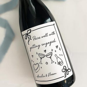 May include: A dark burgundy wine bottle with a white label. The label features the text "Pairs well with getting engaged" and illustrations of champagne glasses, hearts, and a cake. The label is 3.5" x 4".
