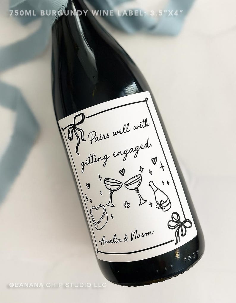 May include: A dark burgundy wine bottle with a white label. The label features the text "Pairs well with getting engaged" and illustrations of champagne glasses, hearts, and a cake. The label is 3.5" x 4".