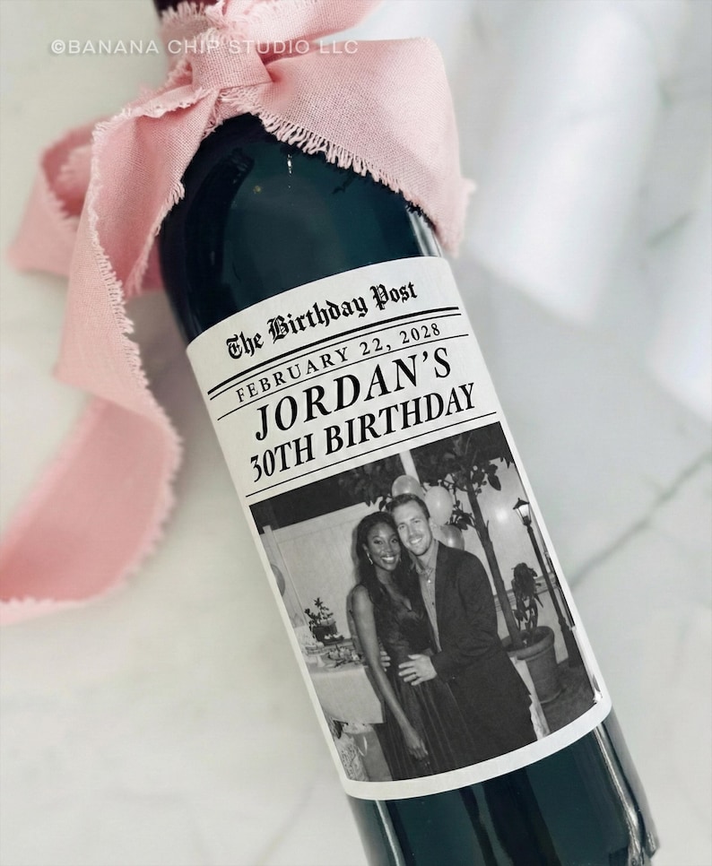 May include: A dark green wine bottle with a custom label that reads "The Birthday Post" and "Jordan's 30th Birthday." The label includes a black and white photo of a couple. A pink ribbon is tied around the bottle's neck.
