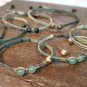 May include: Six green and brown braided bracelets with green and brown beads. The bracelets are on a wooden surface.