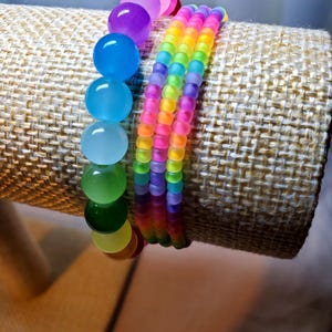 May include: Two beaded bracelets. One bracelet features large, round, translucent beads in a rainbow of colors. The other bracelet has smaller, colorful beads in a rainbow pattern. The bracelets are displayed on a tan, textured surface.